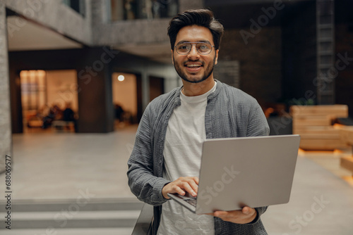 Stylish businessman with laptop standing on modern office background 