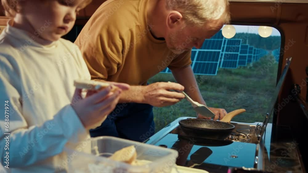 A boy eats a sandwich in a camper as the man cooks beside him, both enjoying the evening at the solar farm A boy eats a sandwich in a camper as the man cooks beside him, both enjoying the evening at the solar farm