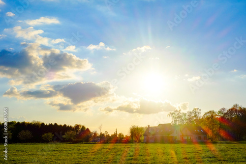 This image captures the serene beauty of a countryside sunset, with the sun's golden rays piercing through the clouds and casting a warm glow over the expansive field. The rays create a dramatic flare photo