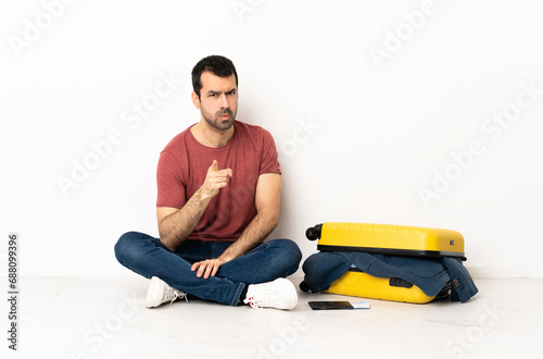 Caucasian handsome man with a suitcase full of clothes sitting on the floor at indoors frustrated and pointing to the front