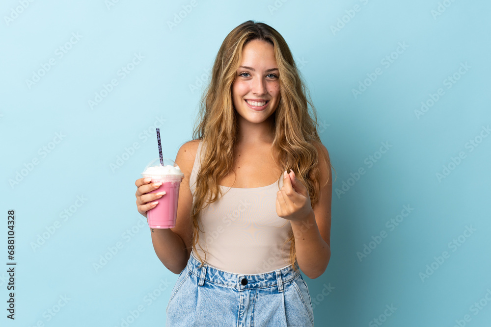 Young woman with strawberry milkshake isolated on blue background making money gesture Young woman with strawberry milkshake isolated on blue background making money gesture