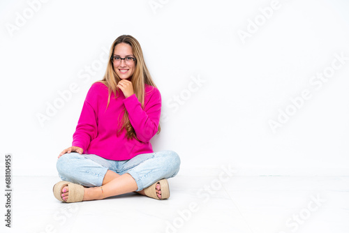 Young caucasian woman sitting on the floor isolated on white background with glasses and smiling