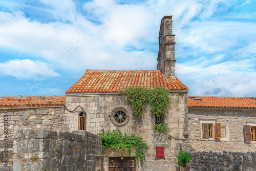 Exterior view of the Church of Santa Maria in Punta in Budva Old Town, Montenegro. Facade of an ancient stone building with a red tiled roof and a round window. Flowering caper bushes grow on the wall