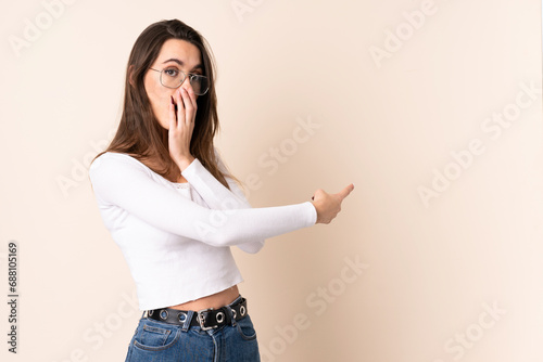 Young woman over isolated beige background with surprise expression while pointing side