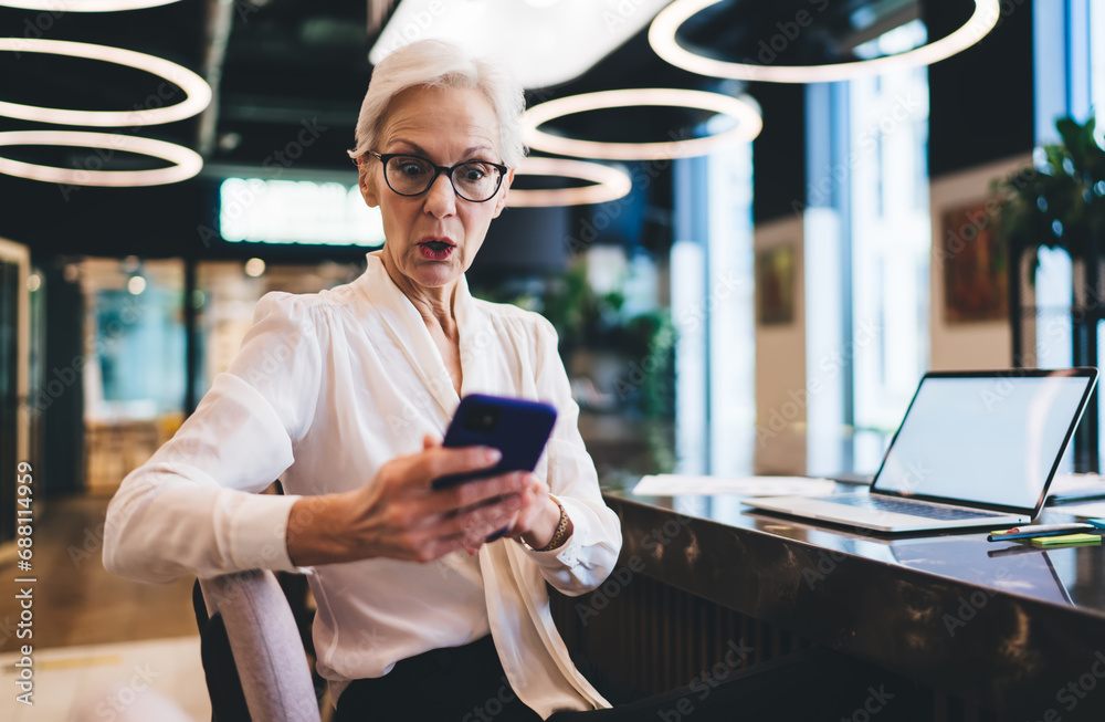 Uncertain senior woman with smartphone sitting at table in office