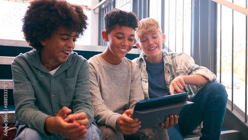 Three Male Secondary Or High School Pupils Inside School Building On Stairs With Digital Tablet