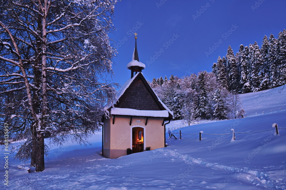 Kapelle zur blauen Stunde an Winterabend bei Neuschnee und Licht aus der Türe