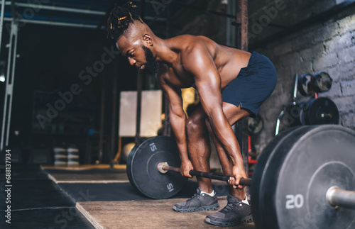 Strong black man bending while lifting heavy barbell in gym