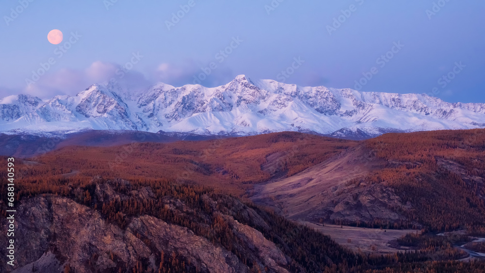 Fototapeta premium Dawn in the mountains with a full moon in the sky. Impressive panorama of dawn snow-capped peaks and a wide mountain valley in soft purple tones.
