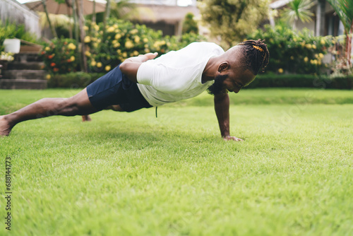 Happy black man performing one arm push up on meadow in garden