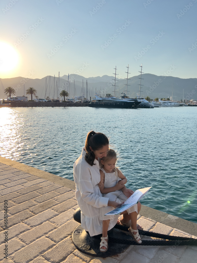 Mom reads a book to a little girl sitting on a bollard against the background of the marina