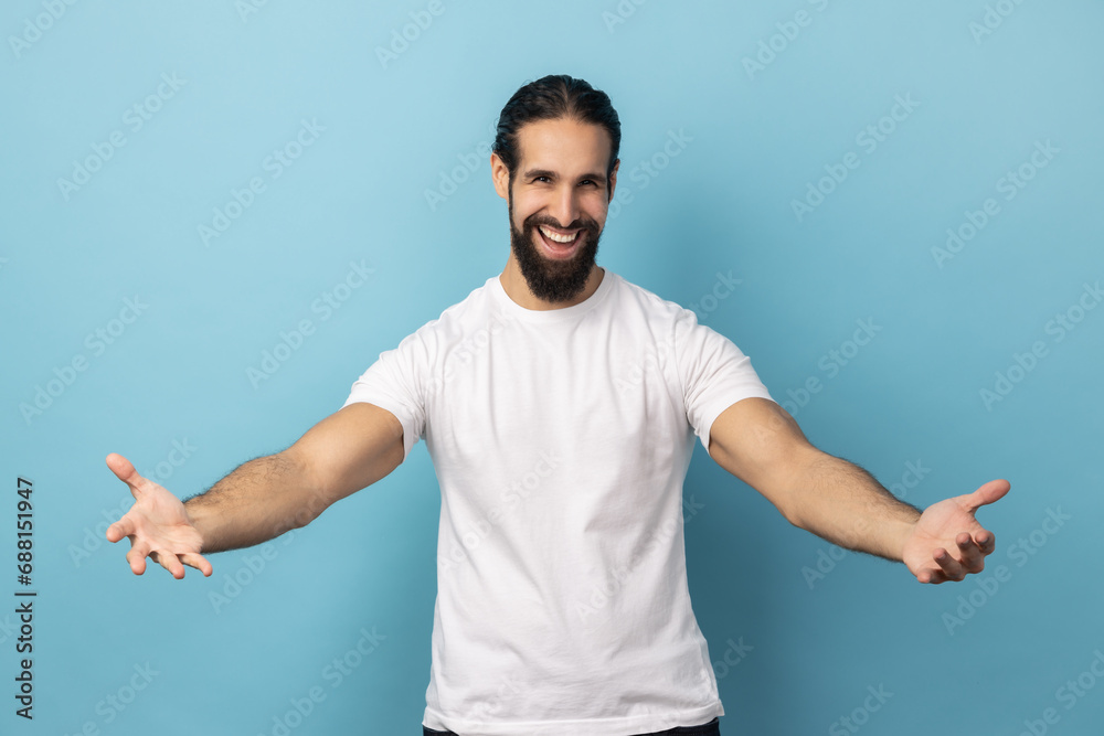 Come into my arms. Portrait of extremely happy man with beard wearing white T-shirt outstretching hands to embrace, giving free hugs and welcoming. Indoor studio shot isolated on blue background. Come into my arms. Portrait of extremely happy man with beard wearing white T-shirt outstretching hands to embrace, giving free hugs and welcoming. Indoor studio shot isolated on blue background.