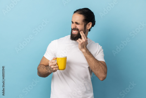 Portrait of man with beard wearing white T-shirt standing and touching his cheek because feeling pain on tooth, after drinking cold water. Indoor studio shot isolated on blue background. photo