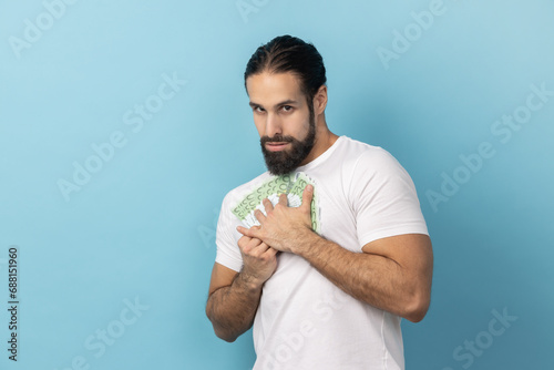 Portrait of man wearing white T-shirt embracing euro banknotes looking at camera with greedy expression, afraid to lose money, obsessed with finances. Indoor studio shot isolated on blue background.