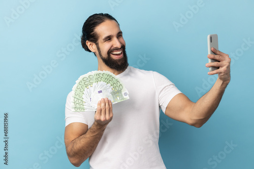 Foto Portrait of delighted optimistic man with beard wearing white T-shirt taking selfie on cellphone with fan of euro banknotes in hands