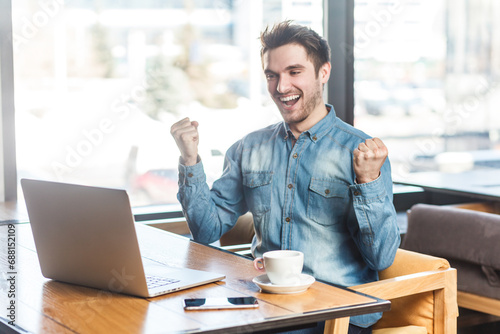 Portrait of extremely happy positive young man freelancer in blue jeans shirt working on laptop, clenched fists, celebrating his success. Indoor shot near big window, cafe background.