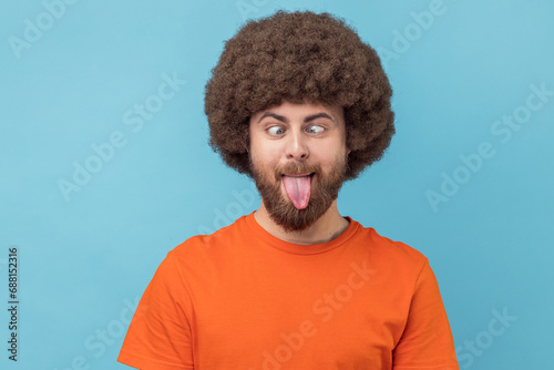 Portrait of silly man with Afro hairstyle wearing orange T-shirt making grimace and crosses eyes, playing fool, having fun alone, sticking out tongue. Indoor studio shot isolated on blue background. photo