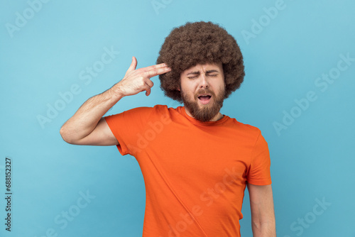 Portrait of frustrated anxious man with Afro hairstyle keeps eyes closed, holding fingers gun pointed to head, shooting himself with hand pistol. Indoor studio shot isolated on blue background.
