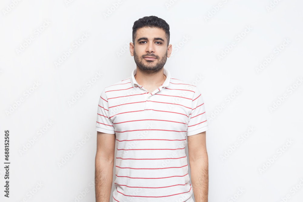 Portrait of strict serious bossy bearded man wearing striped t-shirt standing looking at camera, having assertive facial expression. Indoor studio shot isolated on gray background. Portrait of strict serious bossy bearded man wearing striped t-shirt standing looking at camera, having assertive facial expression. Indoor studio shot isolated on gray background.
