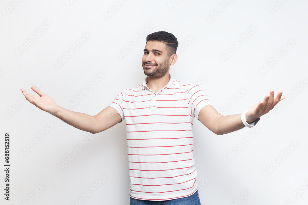 Portrait of friendly kind positive optimistic handsome bearded man wearing striped t-shirt standing with spread hands, inviting guests. Indoor studio shot isolated on gray background. Portrait of friendly kind positive optimistic handsome bearded man wearing striped t-shirt standing with spread hands, inviting guests. Indoor studio shot isolated on gray background.