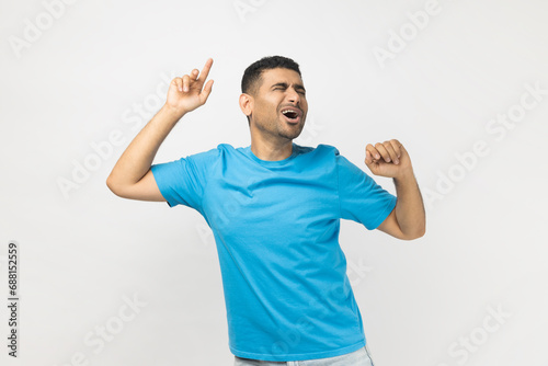 Portrait of happy excited optimistic unshaven man wearing blue T- shirt standing with raised arms, dancing and singing, celebrating holiday. Indoor studio shot isolated on gray background. photo