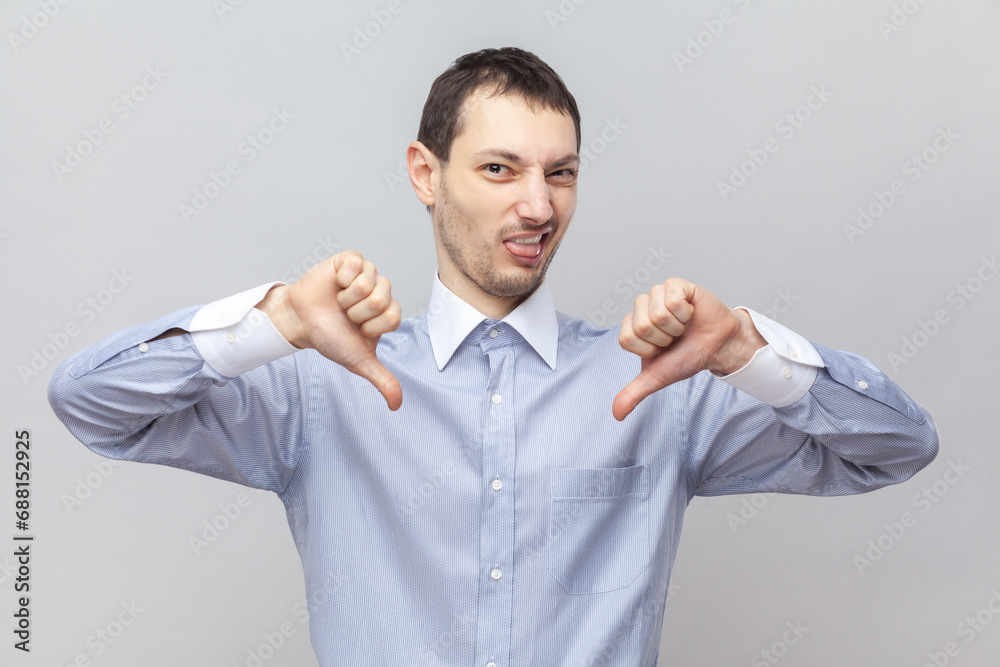 Portrait of disgusted disappointed handsome man standing showing thumbs down, demonstrates dislike gesture, wearing light blue shirt. Indoor studio shot isolated on gray background. Portrait of disgusted disappointed handsome man standing showing thumbs down, demonstrates dislike gesture, wearing light blue shirt. Indoor studio shot isolated on gray background.
