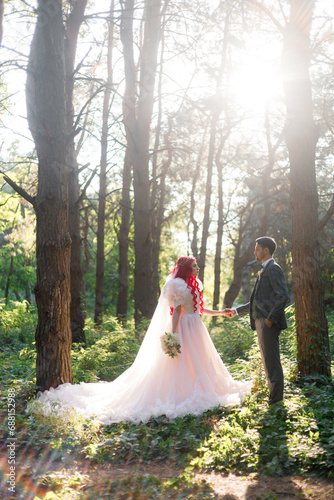 Beautiful couple, the groom in a green suit and the bride in an unusual pink wedding dress, posing at a luxurious ceremony with beautiful decor and an arch with a chandelier and candles in the forest.
