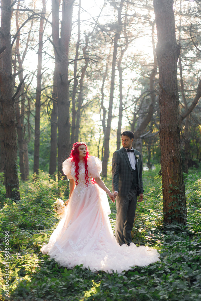 Beautiful couple, the groom in a green suit and the bride in an unusual pink wedding dress, posing at a luxurious ceremony with beautiful decor and an arch with a chandelier and candles in the forest. Beautiful couple, the groom in a green suit and the bride in an unusual pink wedding dress, posing at a luxurious ceremony with beautiful decor and an arch with a chandelier and candles in the forest.