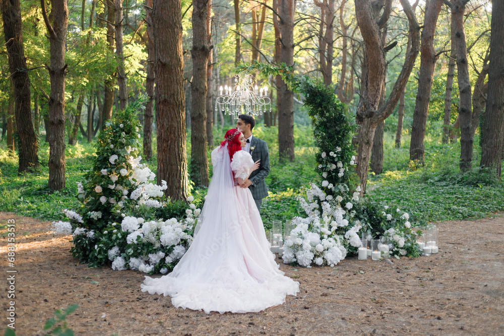 Beautiful couple, the groom in a green suit and the bride in an unusual pink wedding dress, posing at a luxurious ceremony with beautiful decor and an arch with a chandelier and candles in the forest. Beautiful couple, the groom in a green suit and the bride in an unusual pink wedding dress, posing at a luxurious ceremony with beautiful decor and an arch with a chandelier and candles in the forest.