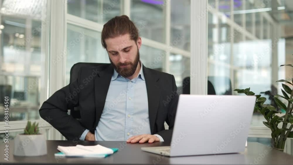 Front view of overtired caucasian businessman working with wireless laptop in own cabinet. Exhausted male dressed in formal suit feeling severe back pain while sitting on office chair indoors. Front view of overtired caucasian businessman working with wireless laptop in own cabinet. Exhausted male dressed in formal suit feeling severe back pain while sitting on office chair indoors.