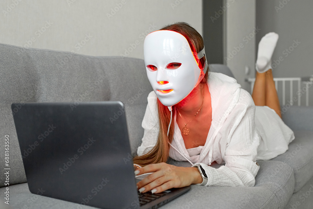 A beautiful girl with an LED mask on her head works at home on a laptop. Home skin care concept.