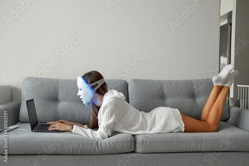 A beautiful girl with an LED mask on her head works at home on a laptop. Home skin care concept.