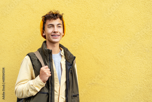 Attractive smiling school boy 
with dental braces on teeth wearing stylish yellow hat, holding backpack  looking away isolated on yellow background, copy space. Education, back to school concept photo