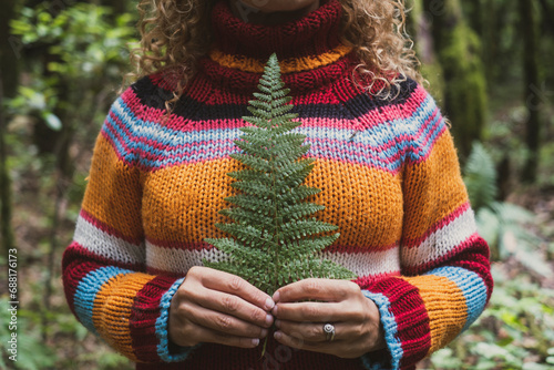 Cut out portrait of unrecognizable woman in nature wearing colorful sweater and holding little tropical leaf against her heart and body. People in love for nature outdoors. Travel healthy lifestyle