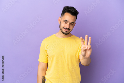 Young caucasian man isolated on purple background happy and counting three with fingers