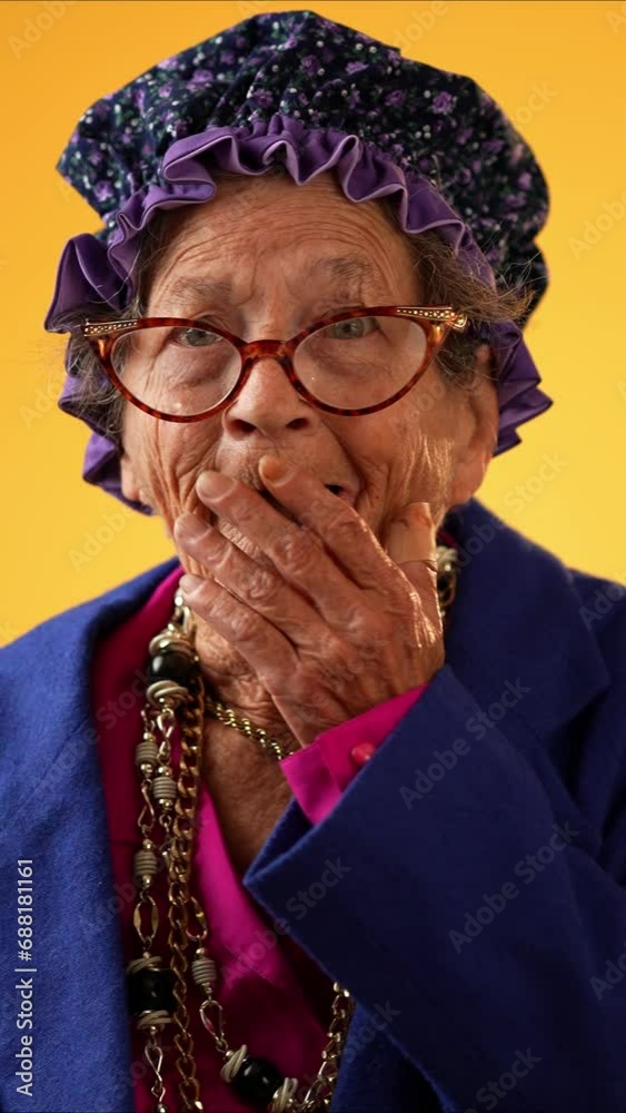 Vertical video portrait of toothless elderly senior old woman with wrinkled skin looks at camera gives mind blown gesture showing explosion of ideas posing isolated on yellow background. 