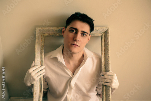 Fotografie Portrait of a handsome young guy in a white shirt, who holds an empty elegant frame from the painting with his hands stained with paint