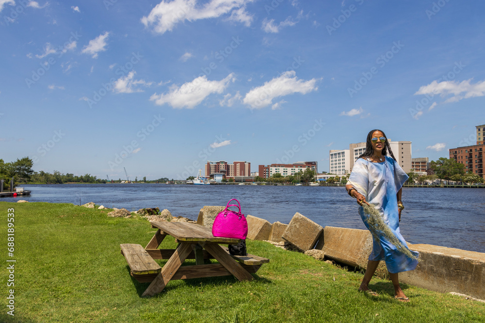 An African American woman with long sisterlocks wearing a blue dress and sunglasses with lush green grass and hotels in the skyline along Cape Fear river in Wilmington North Carolina USA