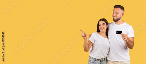 Smiling young european guy and lady in white t-shirts, with credit card hugs, point finger © Prostock-studio