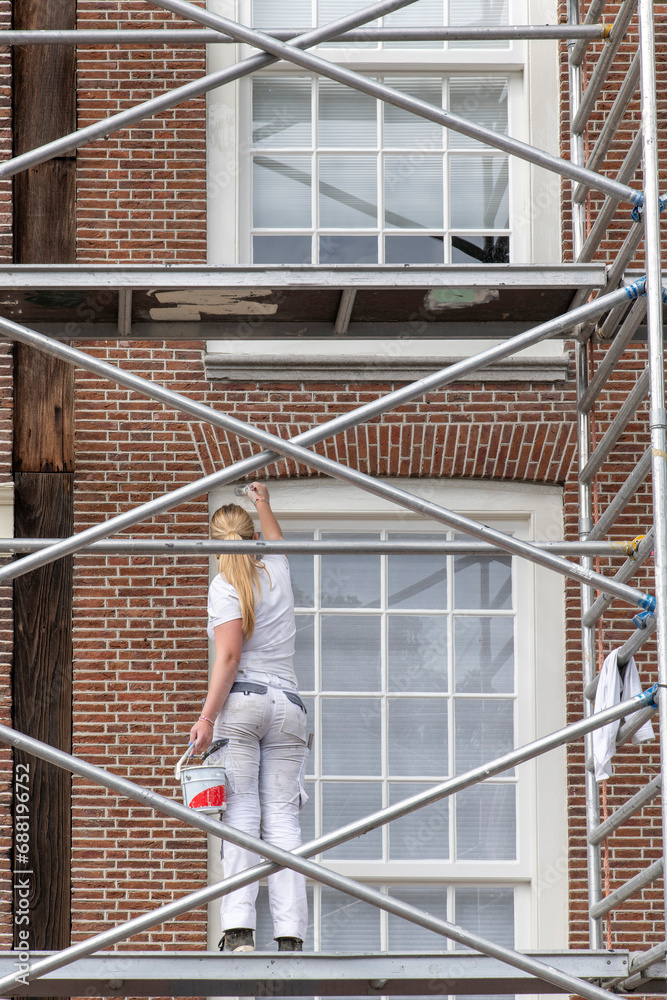Young woman with blond ponytail standing on scaffolding painting white window styles in traditional façade of red brick building; paint bucket in hand Young woman with blond ponytail standing on scaffolding painting white window styles in traditional façade of red brick building; paint bucket in hand