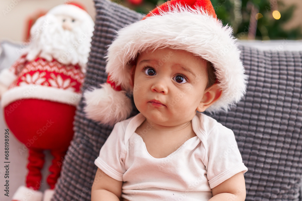 Adorable hispanic baby wearing christmas hat sitting on sofa at home Adorable hispanic baby wearing christmas hat sitting on sofa at home