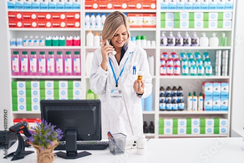Young blonde woman pharmacist talking on telephone holding pills bottle at pharmacy