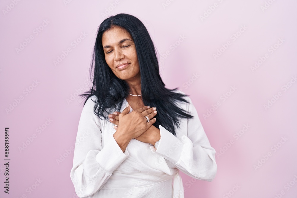 Mature hispanic woman standing over pink background smiling with hands on chest with closed eyes and grateful gesture on face. health concept. Mature hispanic woman standing over pink background smiling with hands on chest with closed eyes and grateful gesture on face. health concept.