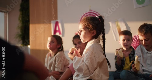 A little girl with a braided hairstyle in a white shirt eats an apple at recess and during lunch in a club for preparing preschool children for school photo