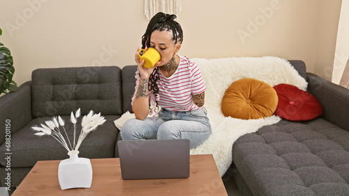 Hispanic amputee woman, seriously focused, utilizing laptop at home; sitting on sofa, sipping coffee, tattoos tracing her armless side. photo