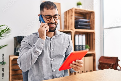 Young hispanic man business worker talking on smartphone using touchpad at office