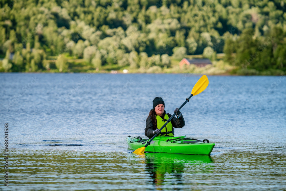 Mature women in green safety life jacket kayaking in green kayak, she looks at camera. Front side photo on still water with blurry green mountain forest background. Sweden.