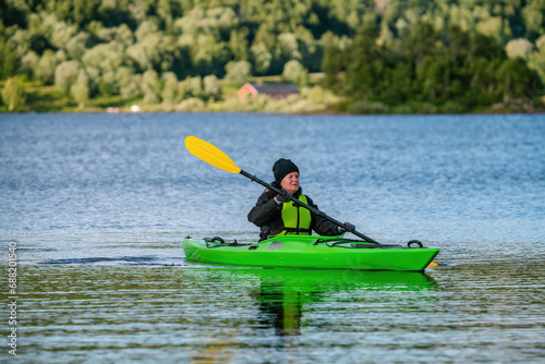 Mature women in green safety life jacket kayaking in green kayak, she looks aside somewhere. Front side photo on still water with blurry green mountain forest background. Sweden.