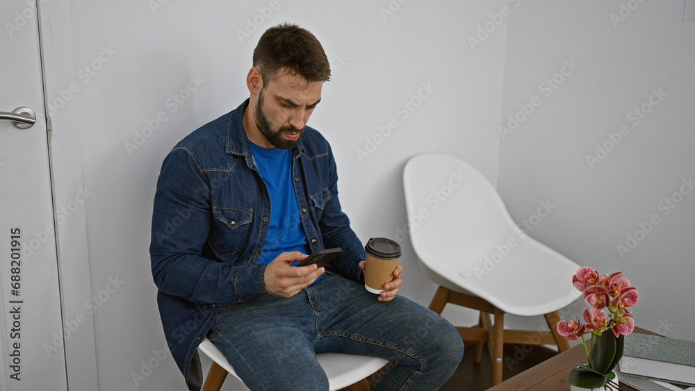 Handsome young hispanic man engrossed texting on smartphone, savoring take away coffee while relaxing in waiting room chair, his concentrated face lit by screen glow.