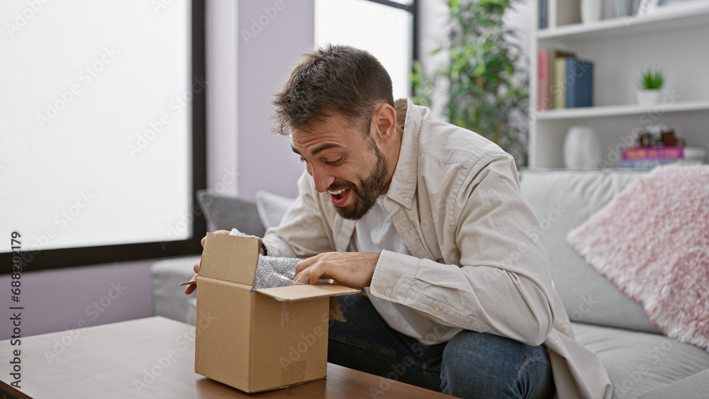 Young, confident hispanic man with a handsome beard happily unpacking a cardboard delivery box while sitting on a sofa at home, enjoying the indoor interior of his apartment living room Young, confident hispanic man with a handsome beard happily unpacking a cardboard delivery box while sitting on a sofa at home, enjoying the indoor interior of his apartment living room